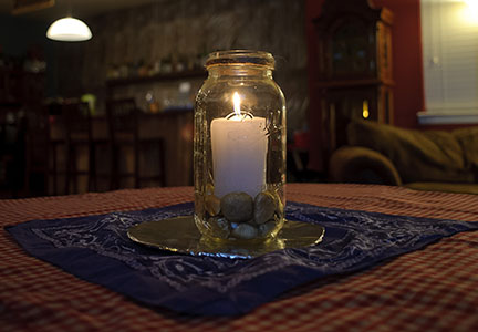 Mason Jar Candle Centerpiece Mason jar with a lit candle and pebbles inside, set on a blue bandana atop a red gingham tablecloth.