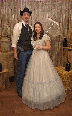 Old West Couple in Murder Mystery Scene Couple dressed in Old West attire posing in a murder mystery setting with rustic props like hay bales and barrels.