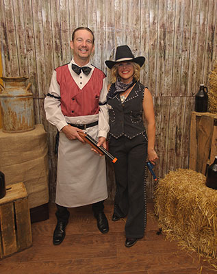 Old West Characters in Rustic Murder Mystery Scene Two people dressed in Old West costumes, holding toy guns, posing in a rustic setting with hay bales and barrels.