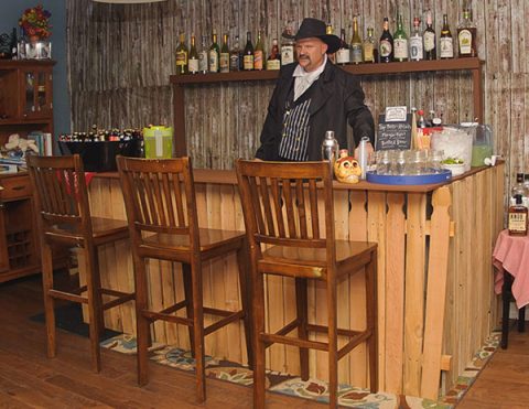 Man dressed in Old West attire standing behind a saloon bar with bottles of liquor and barstools in the foreground.