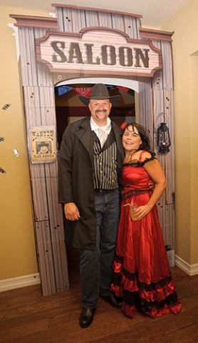 Old West Costumes at Saloon Entrance Man and woman in Old West costumes posing at a saloon-themed entrance with a 'WANTED' poster on the wall.